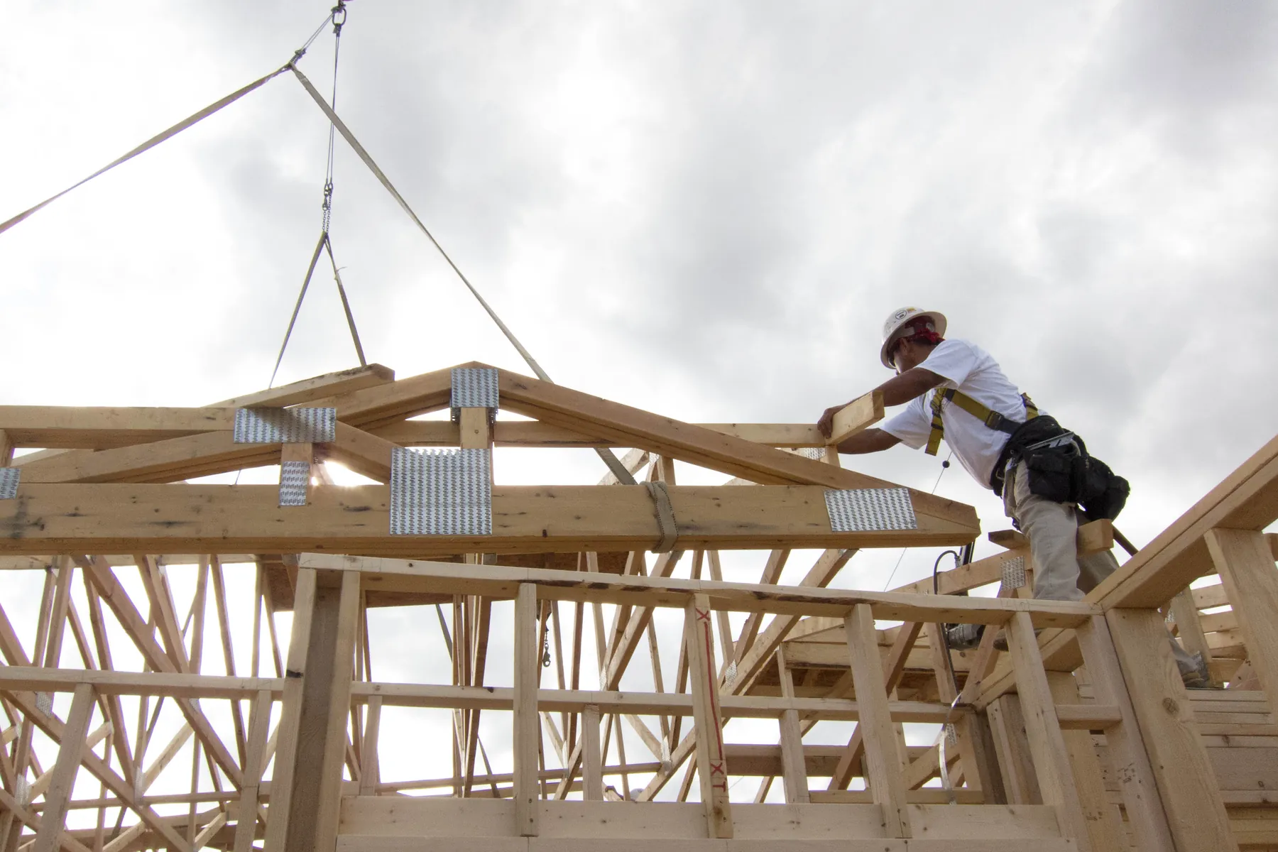 A framer in a fall-protection harness guides a crane-lifted roof truss onto a house under construction