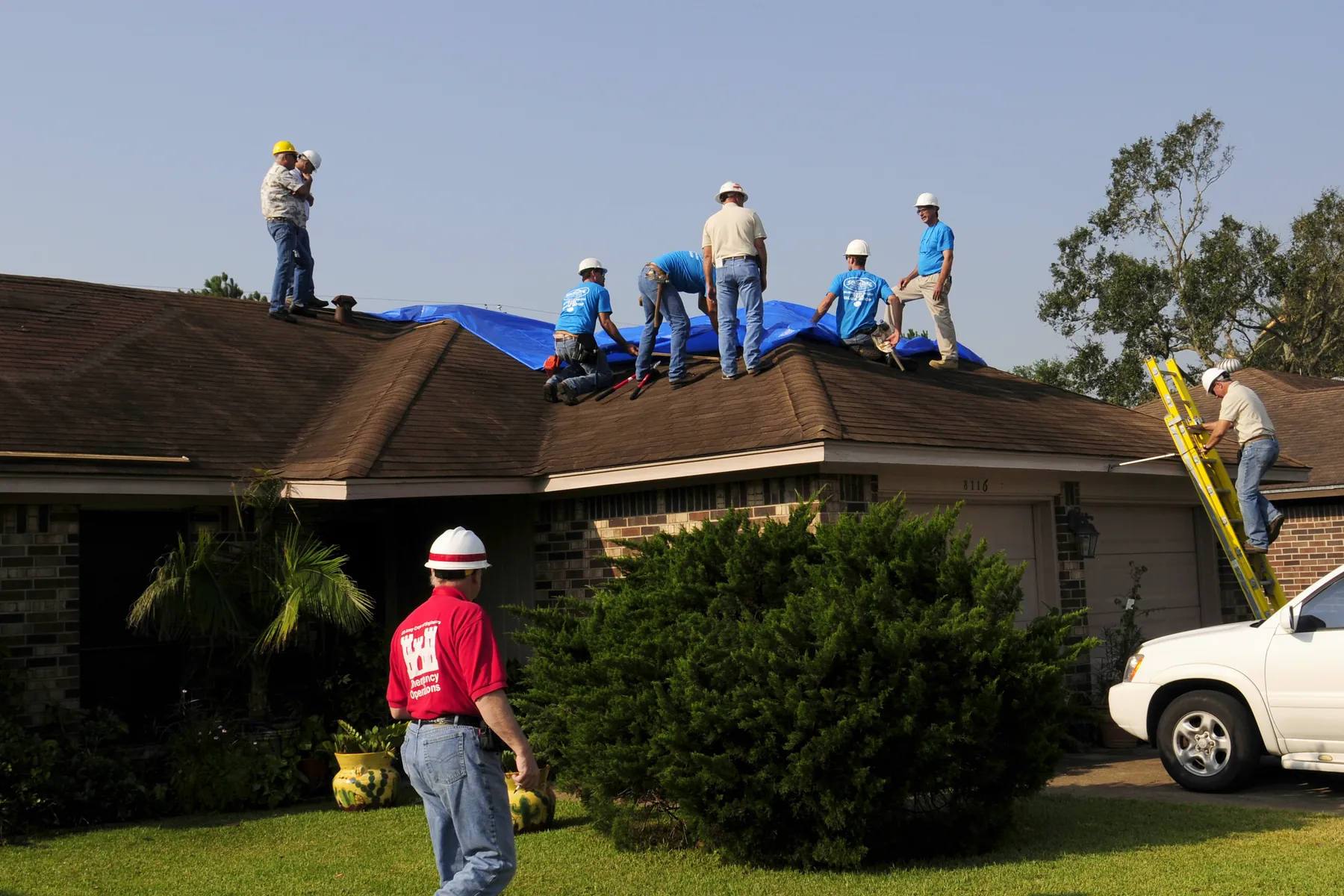 A crew on a storm-damaged house roof spreading a blue tarp while a supervisor walks the yard below