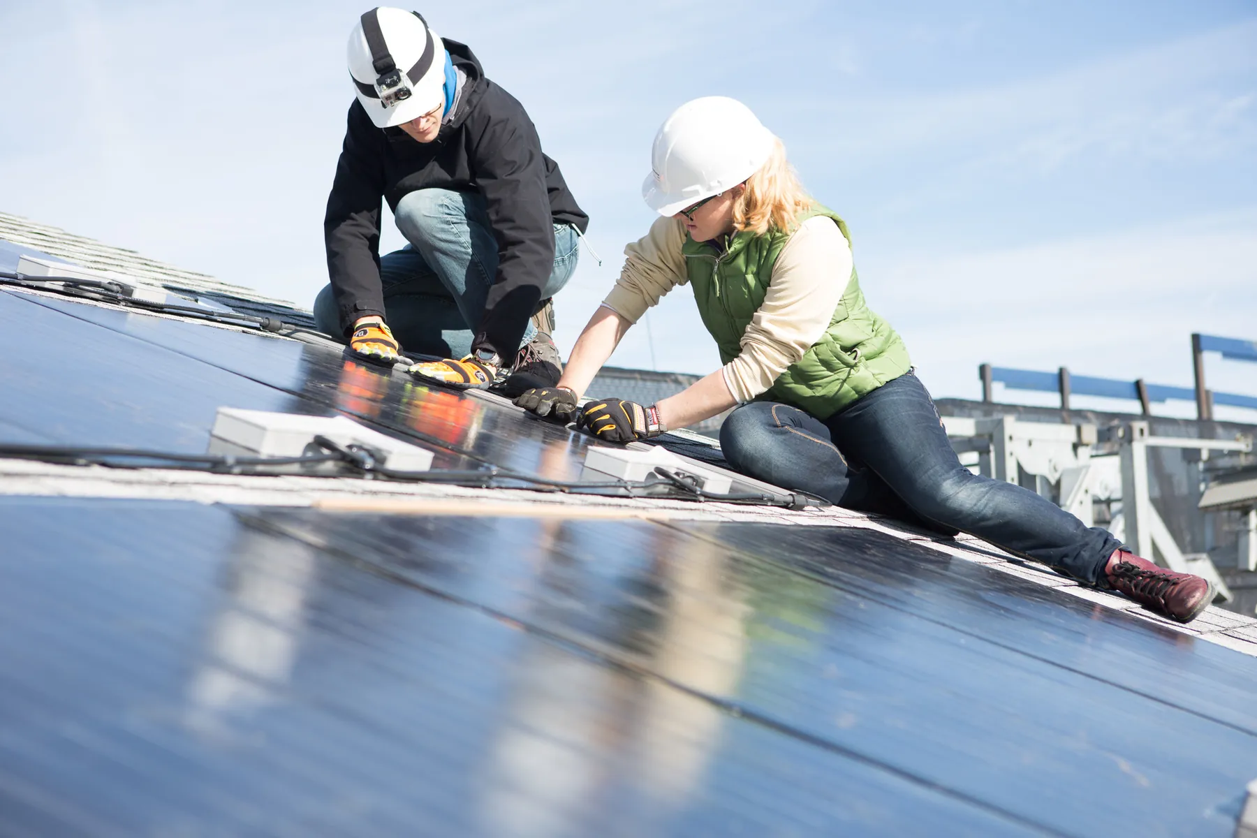 Two installers in hard hats fit solar panels onto a shingled roof